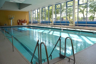 einstieg-ins-becken Ein lichtdurchflutetes Hallenbad mit einem hellblauen Schwimmbecken, das durch ein Geländer leicht zugänglich ist.