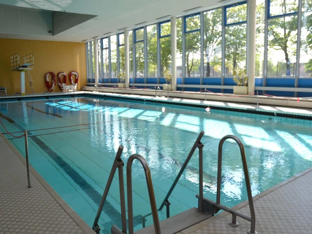 einstieg-ins-becken Ein lichtdurchflutetes Hallenbad mit einem hellblauen Schwimmbecken, das durch ein Geländer leicht zugänglich ist.