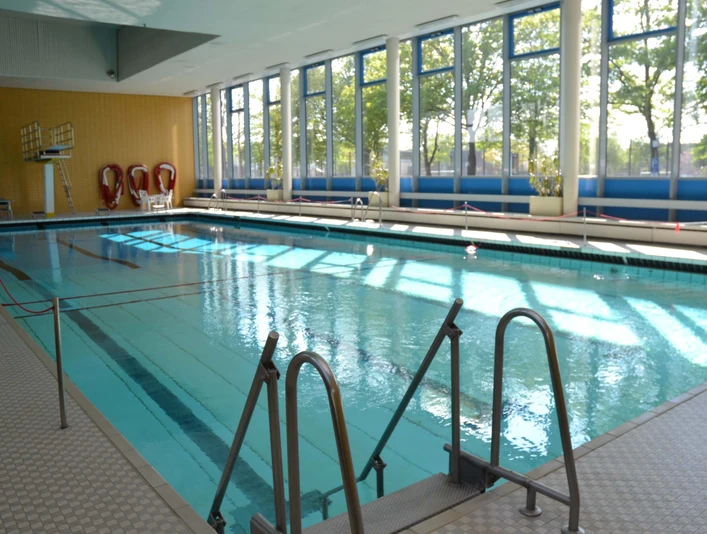 einstieg-ins-becken Ein lichtdurchflutetes Hallenbad mit einem hellblauen Schwimmbecken, das durch ein Geländer leicht zugänglich ist.