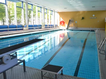 sprungbrett-und-rutsche Ein Hallenbad mit blau gefliestem Becken, Sprungbrett und roter Rutsche, beleuchtet durch große Fenster.