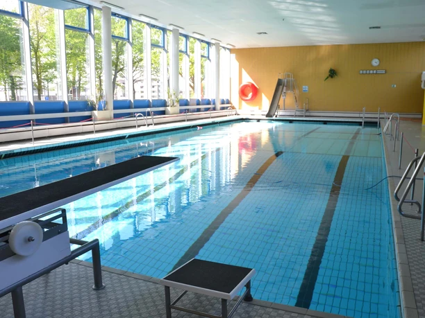 sprungbrett-und-rutsche Ein Hallenbad mit blau gefliestem Becken, Sprungbrett und roter Rutsche, beleuchtet durch große Fenster.