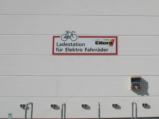E-Bike Ladestation_Möbel Eilers_Apen An einer weißen Wand mit rechteckigen Platten ist ein Schild montiert, das eine Ladestation für Elektro-Fahrräder mit dem Logo von Möbel Eilers in Apen anzeigt.