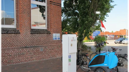 E-Bike Ladestation_Rathausmarkt_Apen Beim Betrachteten Bild handelt es sich um eine E-Bike-Ladestation am Rathausmarkt in Apen. Vor einem Backsteingebäude, die Aper Grundschule, befindet sich eine E-Bike-Ladesäule. Neben der Ladesäule stehen einige Fahrräder, darunter ein Fahrradanhänger in Blau. Im Hintergrund sind weitere Gebäude und parkende Fahrzeuge zu erkennen. Die Umgebung vermittelt ein gepflegtes, verkehrsberuhigtes Bereichsgefühl.E-Bike Ladestation vor einem Backsteingebäude am Rathausmarkt in Apen mit Fahrrädern und Anhänger.