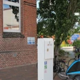 E-Bike Ladestation_Rathausmarkt_Apen Beim Betrachteten Bild handelt es sich um eine E-Bike-Ladestation am Rathausmarkt in Apen. Vor einem Backsteingebäude, die Aper Grundschule, befindet sich eine E-Bike-Ladesäule. Neben der Ladesäule stehen einige Fahrräder, darunter ein Fahrradanhänger in Blau. Im Hintergrund sind weitere Gebäude und parkende Fahrzeuge zu erkennen. Die Umgebung vermittelt ein gepflegtes, verkehrsberuhigtes Bereichsgefühl.E-Bike Ladestation vor einem Backsteingebäude am Rathausmarkt in Apen mit Fahrrädern und Anhänger.