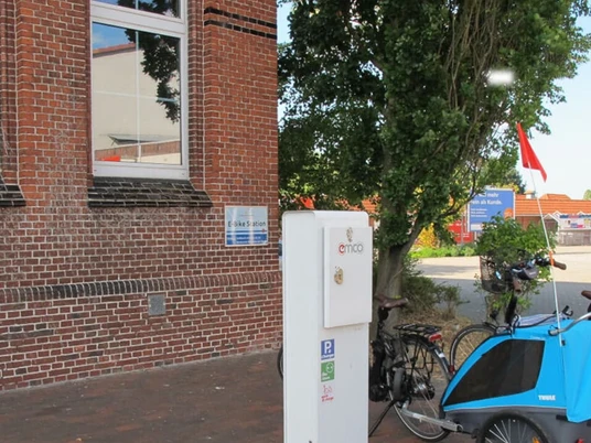 E-Bike Ladestation_Rathausmarkt_Apen Beim Betrachteten Bild handelt es sich um eine E-Bike-Ladestation am Rathausmarkt in Apen. Vor einem Backsteingebäude, die Aper Grundschule, befindet sich eine E-Bike-Ladesäule. Neben der Ladesäule stehen einige Fahrräder, darunter ein Fahrradanhänger in Blau. Im Hintergrund sind weitere Gebäude und parkende Fahrzeuge zu erkennen. Die Umgebung vermittelt ein gepflegtes, verkehrsberuhigtes Bereichsgefühl.E-Bike Ladestation vor einem Backsteingebäude am Rathausmarkt in Apen mit Fahrrädern und Anhänger.