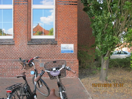Ladestation_Rathausmarkt_Apen Zwei Fahrräder stehen vor einem Backsteingebäude, das mit einem Schild "E-Bike Station" gekennzeichnet ist.