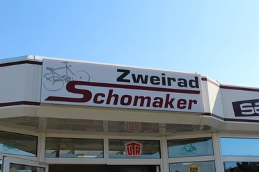 Zweirad Schomaker Eingangsbereich von Zweirad Schomaker mit Firmenschild und Fahrrad-Symbol unter blauem Himmel.