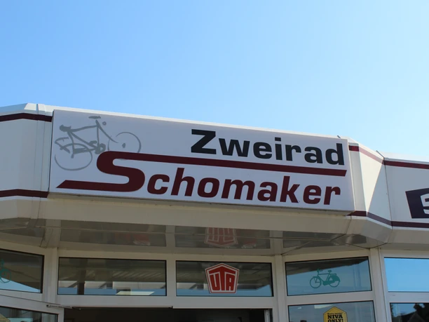 Zweirad Schomaker Eingangsbereich von Zweirad Schomaker mit Firmenschild und Fahrrad-Symbol unter blauem Himmel.