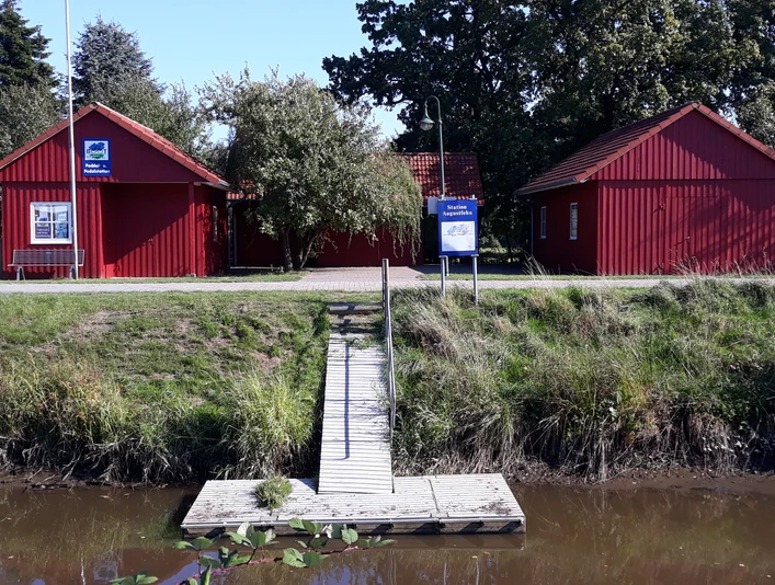 Paddel und Pedal Station Die Paddel- und Pedalstation zeigt zwei rot gestrichene, skandinavische Holzhütten, umgeben von Natur.