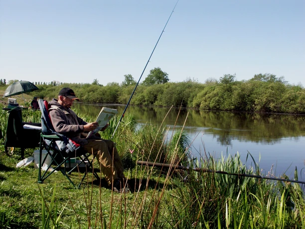 Angeln_Erholungsort Rhauderfehn Mann sitzt auf einem Klappstuhl am Flussufer und liest Zeitung, mehrere Angelruten sind im Wasser platziert