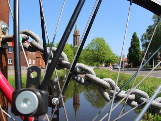Rhauderfehn erfahren - Wiekentour - Gemeinde Rhauderfehn, Ostfriesland, Rad steht auf Fehnbrücke und Blick auf den Rajen mit der Hoffnungskirche im Zentrum Ein Fahrrad mit Blick durch das Hinterrad und im Hindergrund eine Brückenkette, ein Kanal und eine Kirche.