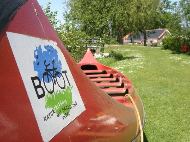 Paddel- und Pedal Rhauderfehn, Ostfriesland Rotes Kanu mit Aufkleber 'BOOT Naturerlebnis mit Paddel und Pedal' auf einer grünen Wiese mit Bäumen und Häusern im Hintergrund