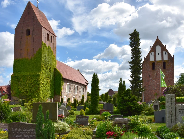 Die St.-Johannes-Kirche erhebt sich majestätisch über den gepflegten Kirchhof mit blühenden Beeten und Grabsteinen. Der Turm ist mit Efeu bewachsen, was ihm ein malerisches Aussehen verleiht.