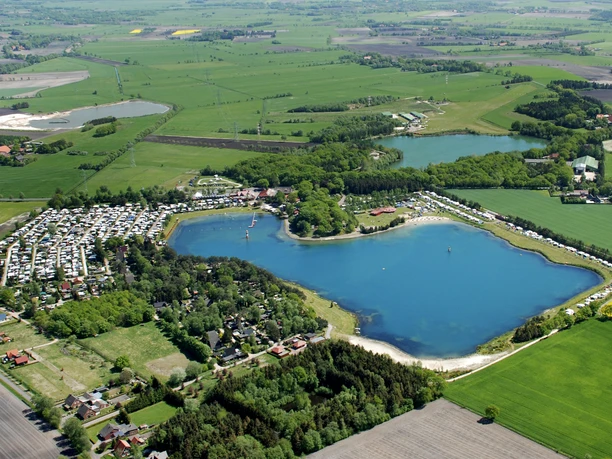 Luftaufnahme Bernsteinsee Die Luftaufnahme zeigt den Bernsteinsee umgeben von grünen Feldern, einem Campingplatz und Bäumen.