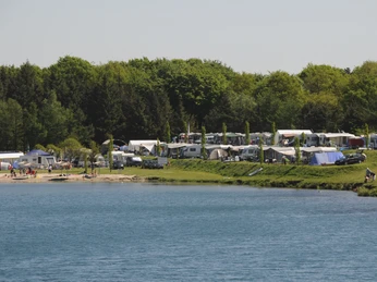 Camping am Bernsteinsee Am Ufer des Bernsteinsees stehen viele Zelte und Wohnwagen, umgeben von Bäumen und grünem Rasen.