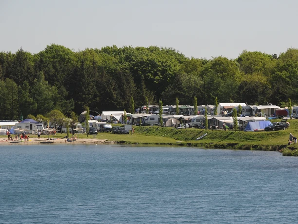 Camping am Bernsteinsee Am Ufer des Bernsteinsees stehen viele Zelte und Wohnwagen, umgeben von Bäumen und grünem Rasen.