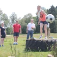 Auf einer grünen Wiese spielt eine Gruppe von vier Menschen Fußballgolf. Eine Frau steht lächelnd auf einem großen Traktorreifen und kickt einen Fußball. Im Hintergrund lachen drei weitere Personen. Der sonnige Tag und die entspannte, spielerische Atmosphäre sind spürbar.