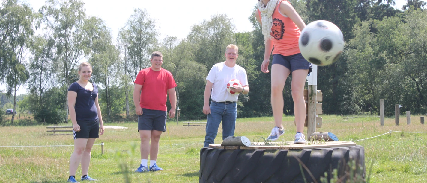 Auf einer grünen Wiese spielt eine Gruppe von vier Menschen Fußballgolf. Eine Frau steht lächelnd auf einem großen Traktorreifen und kickt einen Fußball. Im Hintergrund lachen drei weitere Personen. Der sonnige Tag und die entspannte, spielerische Atmosphäre sind spürbar.