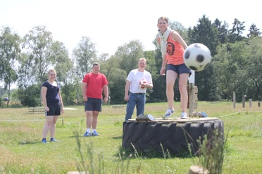 Fußballgolf Auf einer grünen Wiese spielt eine Gruppe von vier Menschen Fußballgolf. Eine Frau steht lächelnd auf einem großen Traktorreifen und kickt einen Fußball. Im Hintergrund lachen drei weitere Personen. Der sonnige Tag und die entspannte, spielerische Atmosphäre sind spürbar.