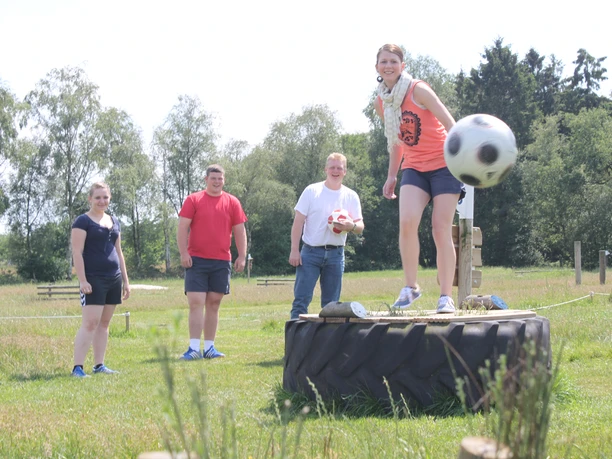 Fußballgolf Auf einer grünen Wiese spielt eine Gruppe von vier Menschen Fußballgolf. Eine Frau steht lächelnd auf einem großen Traktorreifen und kickt einen Fußball. Im Hintergrund lachen drei weitere Personen. Der sonnige Tag und die entspannte, spielerische Atmosphäre sind spürbar.