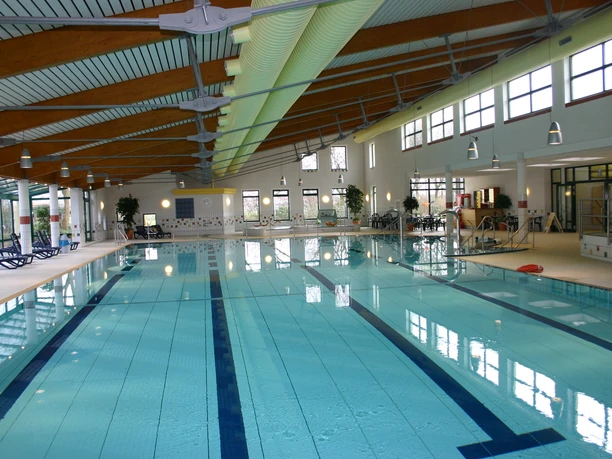 Hallenbad Das Bild zeigt ein modernes Hallenbad mit einem großen Schwimmbecken, umgeben von hellen Fliesen und großen Fenstern, die natürliches Licht einlassen.