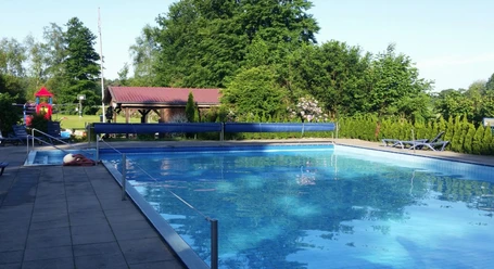 Das Bild zeigt ein Freibad mit einem großen rechteckigen Pool, umgeben von Bäumen und einer Wiese. Im Hintergrund ist ein kleines Gebäude mit rotem Dach zu sehen. Links vom Bild sieht man einen Kinderspielplatz mit bunten Spielgeräten. Diese erholsame Umgebung lädt zum Entspannen und Spielen ein.
