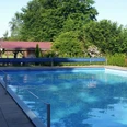 Das Bild zeigt ein Freibad mit einem großen rechteckigen Pool, umgeben von Bäumen und einer Wiese. Im Hintergrund ist ein kleines Gebäude mit rotem Dach zu sehen. Links vom Bild sieht man einen Kinderspielplatz mit bunten Spielgeräten. Diese erholsame Umgebung lädt zum Entspannen und Spielen ein.