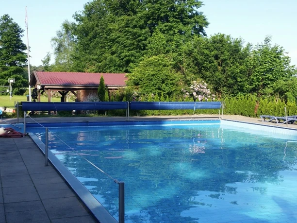 Das Bild zeigt ein Freibad mit einem großen rechteckigen Pool, umgeben von Bäumen und einer Wiese. Im Hintergrund ist ein kleines Gebäude mit rotem Dach zu sehen. Links vom Bild sieht man einen Kinderspielplatz mit bunten Spielgeräten. Diese erholsame Umgebung lädt zum Entspannen und Spielen ein.