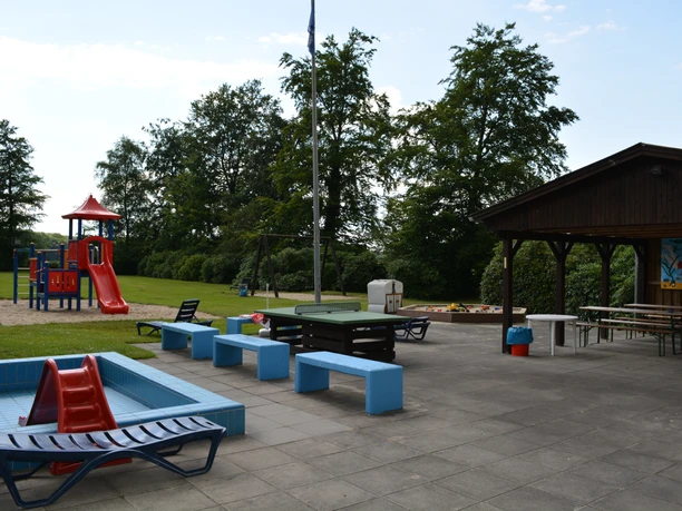 Ein Spielplatz mit Rutsche, Wipp-Tier, Picknickbänken und überdachtem Essbereich in einer grünen Umgebung.