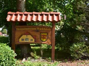 Heimatmuseum-Wiefelstede Holzschild mit Aufschrift 'Heimatmuseum Wiefelstede', umgeben von grünen Bäumen und Pflanzen.