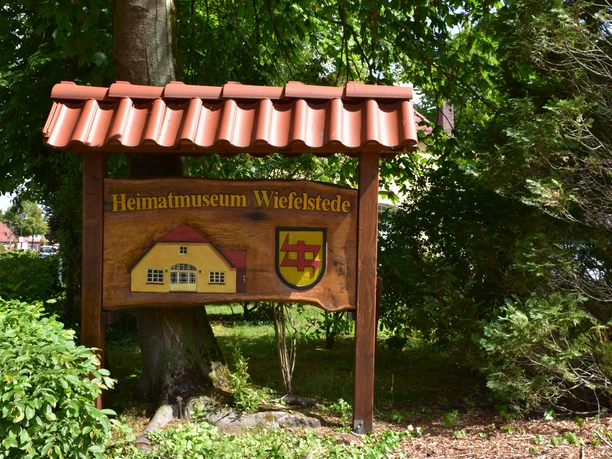 Heimatmuseum-Wiefelstede Holzschild mit Aufschrift 'Heimatmuseum Wiefelstede', umgeben von grünen Bäumen und Pflanzen.