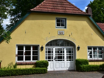 Heimatmuseum-Wiefelstede Gelbes Haus mit rotem Dach, weißen Fenstern und Eingang. Schriftzug „Heimatmuseum“ über der Tür.