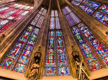 Im Aachener Dom befindet sich ein Ensemble aus hoch aufragenden, bunten Glasfenstern. Die lebendigen Farben der Fenster tauchen den Raum in ein mystisches Lichtspiel, ergänzt durch Skulpturen einiger Heiligen darunter.