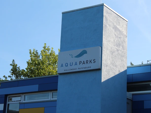 hallenbad-papenburg-02-emsland-tourismus-christina-poll Blauer Gebäudeturm mit dem Schild Aqua Parks Hallenbad Papenburg vor klarem Himmel und Bäumen.