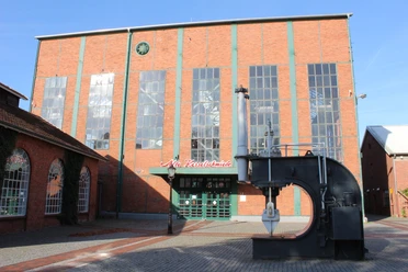 Alte Kesselschmiede Backsteingebäude mit hohen Fenstern und großem Metallhammer vor dem Eingang der Alten Kesselschmiede.