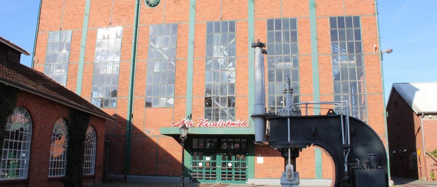 Alte Kesselschmiede Backsteingebäude mit hohen Fenstern und großem Metallhammer vor dem Eingang der Alten Kesselschmiede.