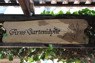 Arns Gartenidyll Holzschild mit der Aufschrift „Arns’ Gartenidylle“ und Blumengravur, umgeben von Efeuranken.