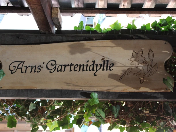 Arns Gartenidyll Holzschild mit der Aufschrift „Arns’ Gartenidylle“ und Blumengravur, umgeben von Efeuranken.