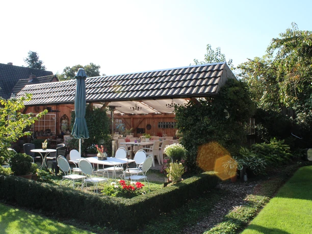 arns-gertenidylle-01-emsland-tourismus-christina-poll Sonniger Garten mit überdachter Terrasse, bepflanzten Wegen und Sitzplätzen im Grünen.