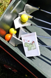 Ausstattung Swingolf Vier Golfschläger und farbige Golfbälle in einem Transportwagen, daneben eine Karte der Golfanlage.