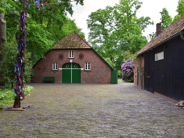 Heinrich Kunst Haus Das Bild zeigt einen kleinen idyllischen Hof umgeben von Bäumen. Im Zentrum steht ein traditionelles rotes Backsteinhaus mit grünem Holztor und kleinen runden Fenstern. Rechts daneben befindet sich ein dunkles Fachwerkgebäude. Die gepflasterte Fläche und das grüne Blattwerk erzeugen eine ruhige, einladende Atmosphäre. Dieses Setting vermittelt den Charme ländlichen Lebens und Ruhe.