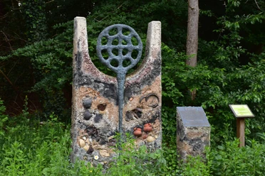 Hügelgrab Ein modernes Kunstwerk aus Stein, markiert und verziert mit Kreisen, Flaschen und kleinen Töpfen, steht vor einem bewaldeten Hintergrund.