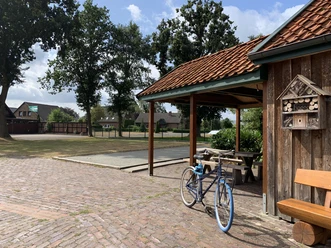 Boule Hof Kleiberg Gepflasterter Hof mit Holzgebäude, kleinem Sitzplatz, Parkbank und blauem Fahrrad. Umgeben von Bäumen und Boulefeld. Ruhiges Ambiente.
