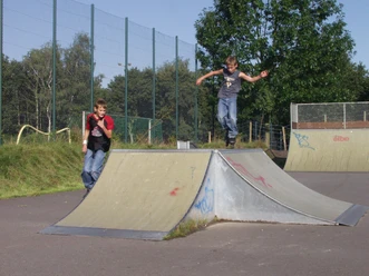 Skatepark Zwei Jungen beim Skaten im Skatepark. Einer springt von einer Rampe, während der andere daneben steht. Umgeben von Grün und einem hohen Zaun im Hintergrund.