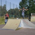 Skatepark Zwei Jungen beim Skaten im Skatepark. Einer springt von einer Rampe, während der andere daneben steht. Umgeben von Grün und einem hohen Zaun im Hintergrund.