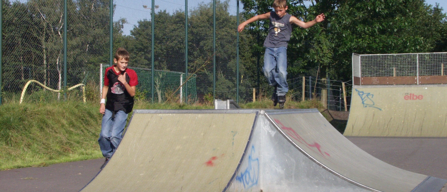 Skatepark Zwei Jungen beim Skaten im Skatepark. Einer springt von einer Rampe, während der andere daneben steht. Umgeben von Grün und einem hohen Zaun im Hintergrund.