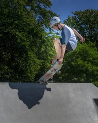 Skatepark Ein Skateboarder mit Helm macht einen Trick auf einer Rampe bei strahlendem Sonnenschein, umgeben von grünen Bäumen.