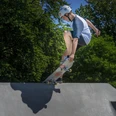 Skatepark Ein Skateboarder mit Helm macht einen Trick auf einer Rampe bei strahlendem Sonnenschein, umgeben von grünen Bäumen.