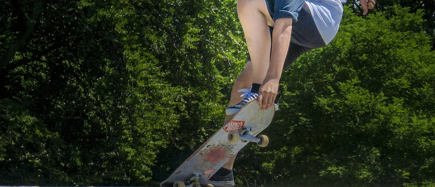 Skatepark Ein Skateboarder mit Helm macht einen Trick auf einer Rampe bei strahlendem Sonnenschein, umgeben von grünen Bäumen.