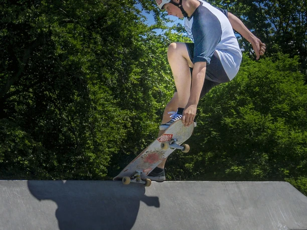Skatepark Ein Skateboarder mit Helm macht einen Trick auf einer Rampe bei strahlendem Sonnenschein, umgeben von grünen Bäumen.
